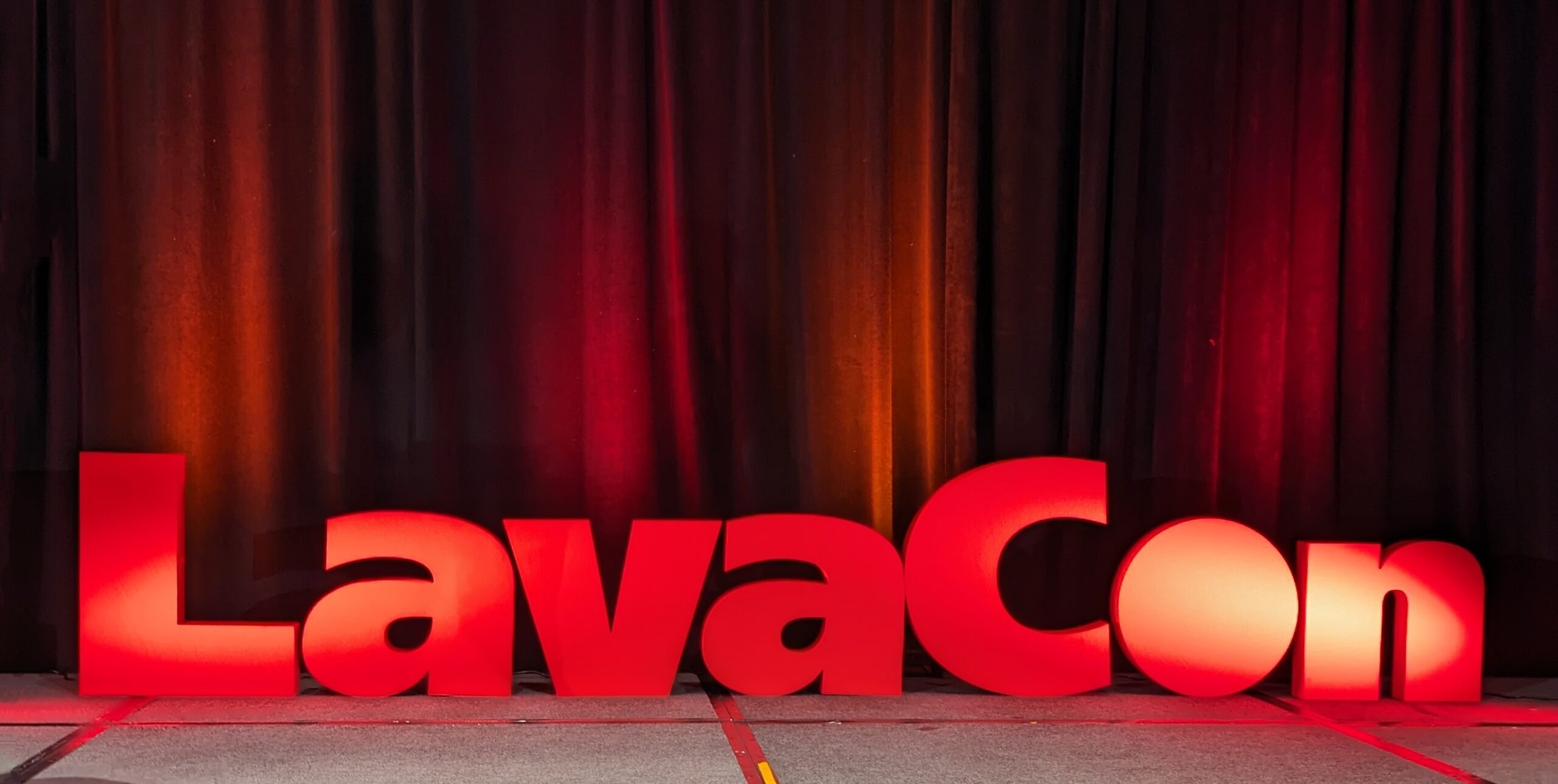 Grey stage and black curtains with large red letters standing up on stage that spell “LavaCon” and are lit up.