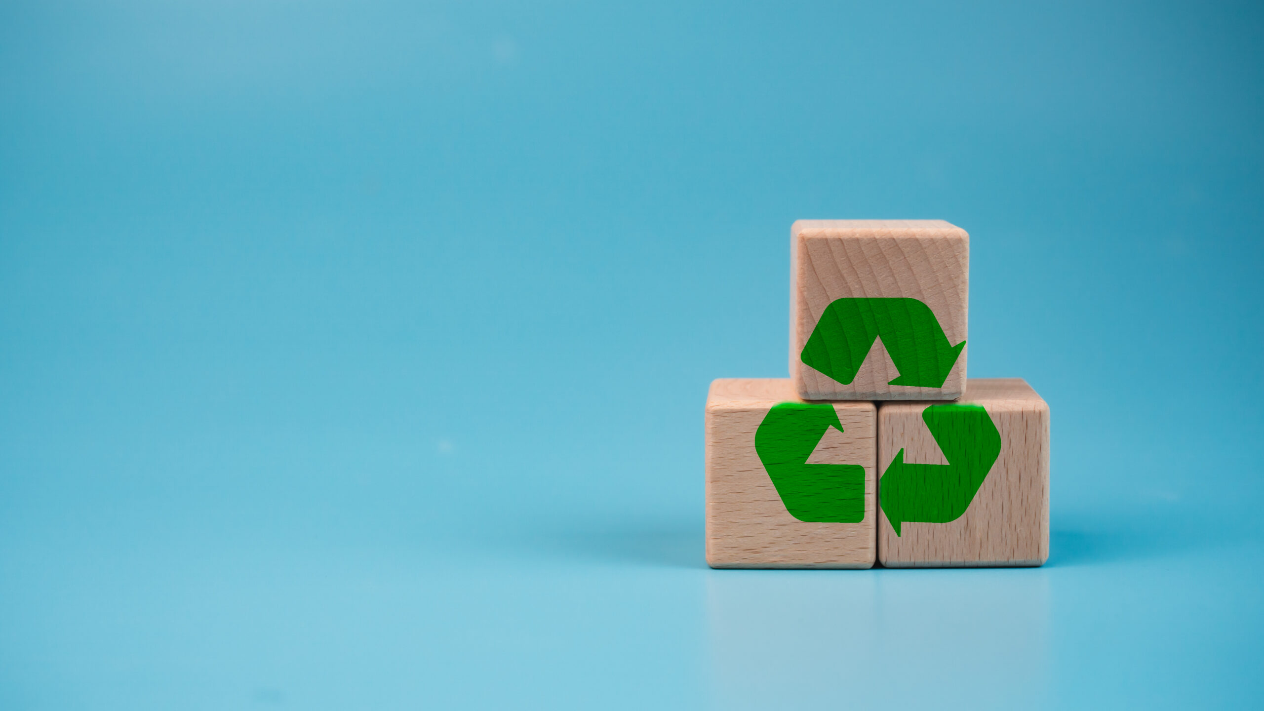 Wooden blocks arranged in a pyramid against a blue background, each displaying a part of a green recycling symbol, forming a complete recycle icon when viewed together.