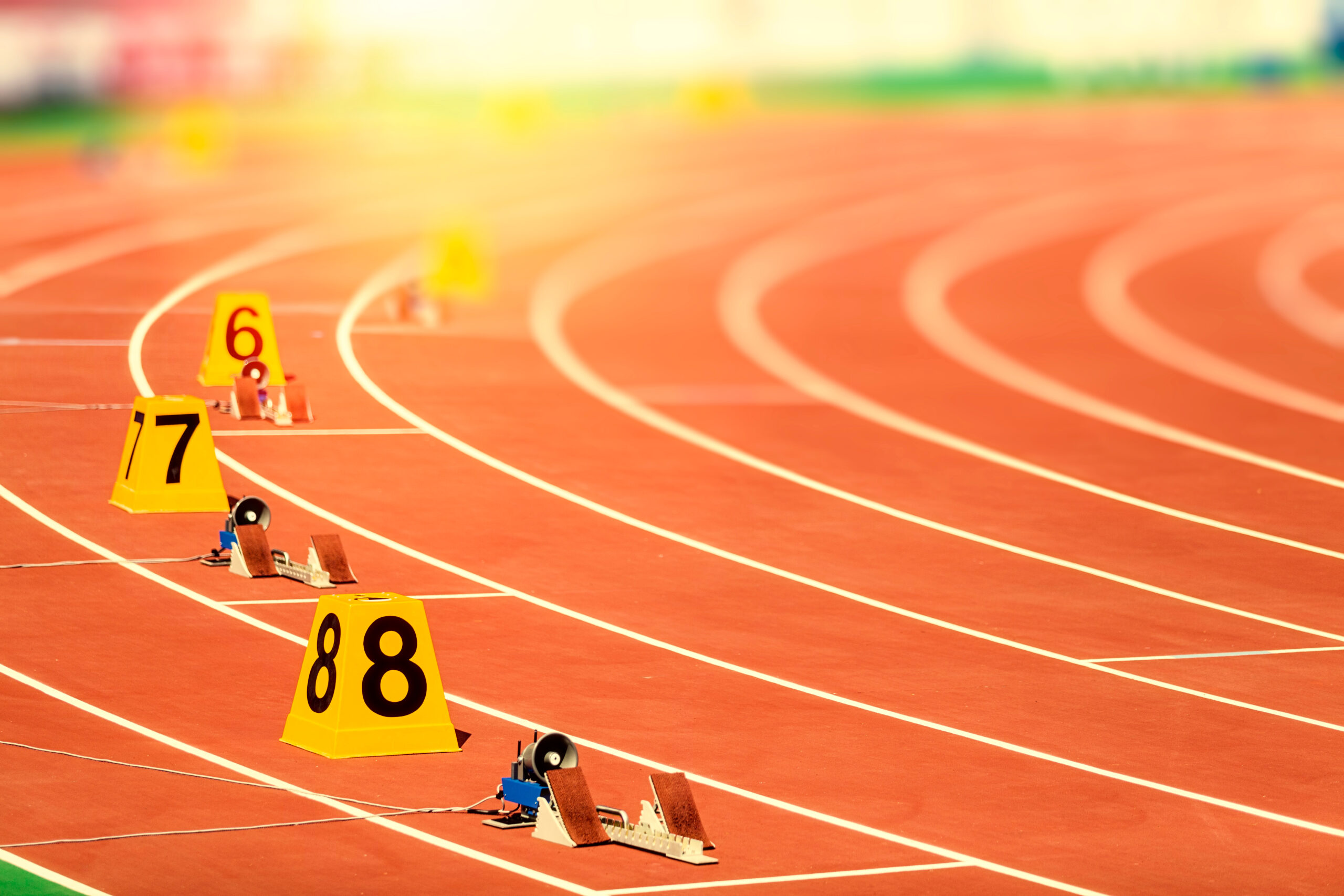 A sunlit running track with multiple lanes, each marked with a yellow cone displaying lane numbers 6, 7, and 8. Starting blocks are set up at the beginning of each lane, ready for a sprint race. The track curves into the distance with a soft blur, suggesting early morning or late afternoon light.