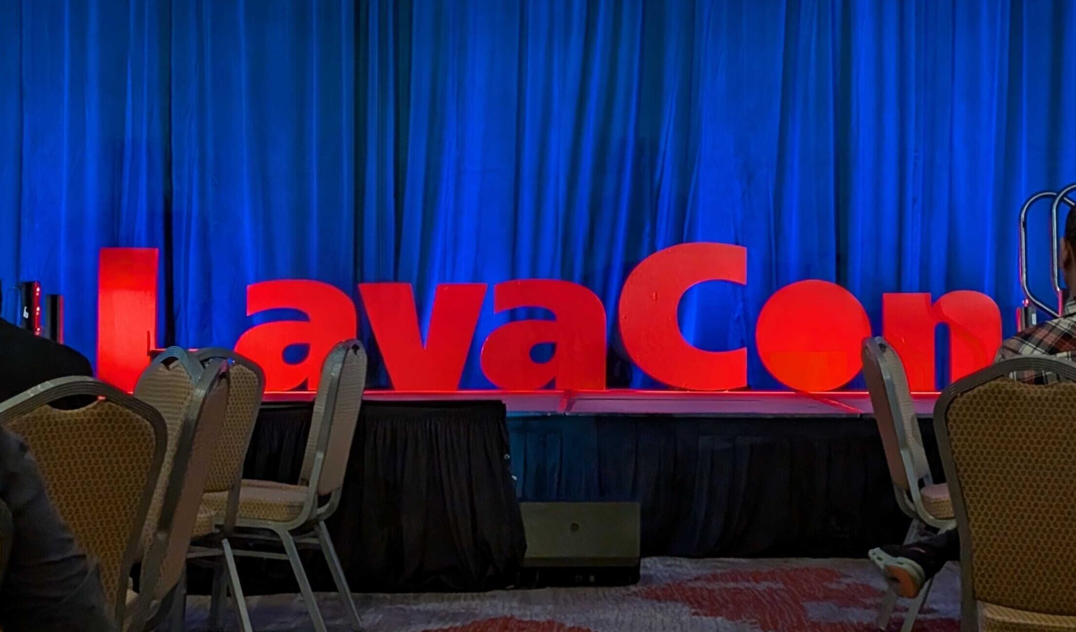 A stage with large red letters spelling “LavaCon” against a blue curtain backdrop. A few empty chairs visible in the foreground.